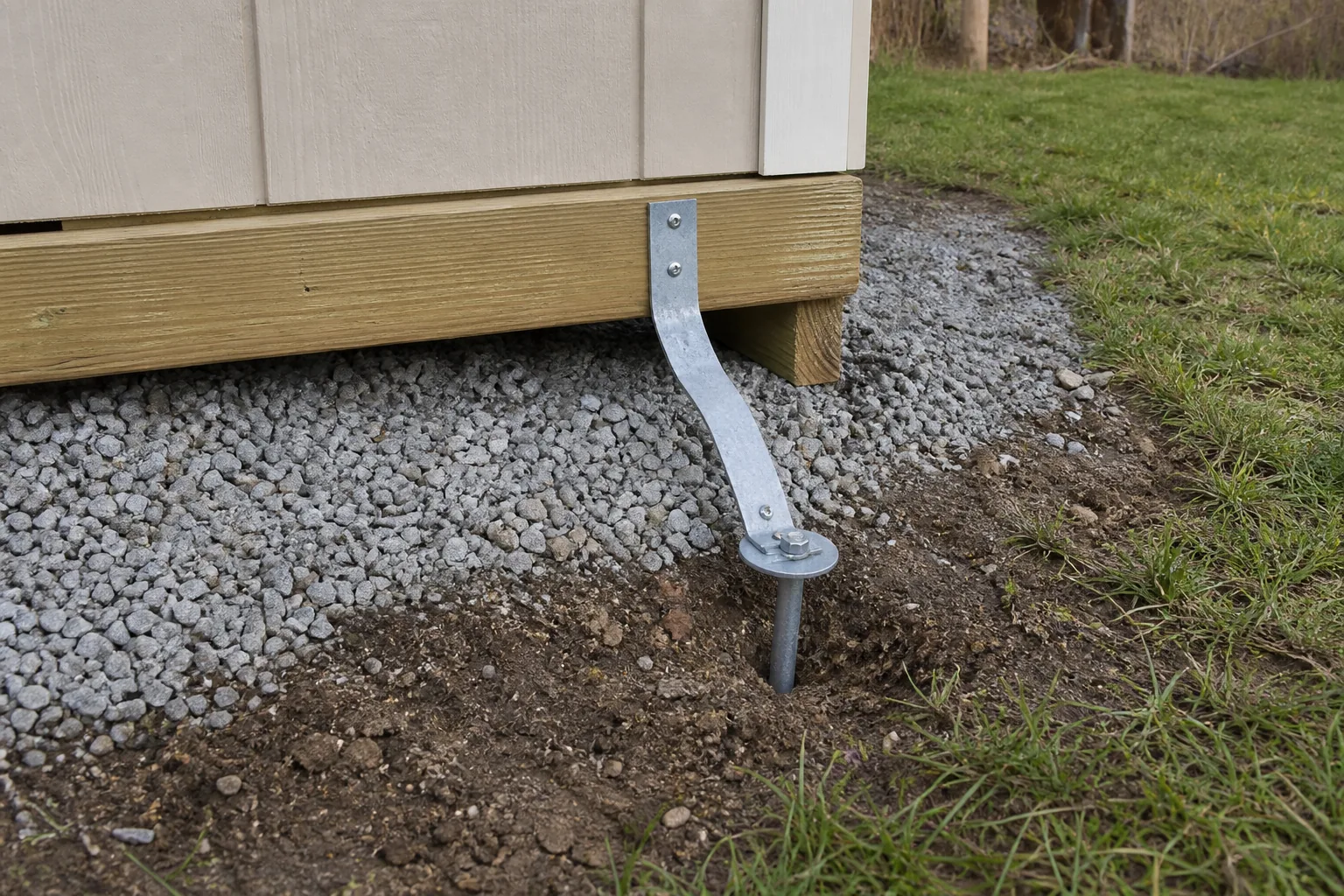 Close view of a structural shed skid connected by a metal strap to a ground anchor in firm soil