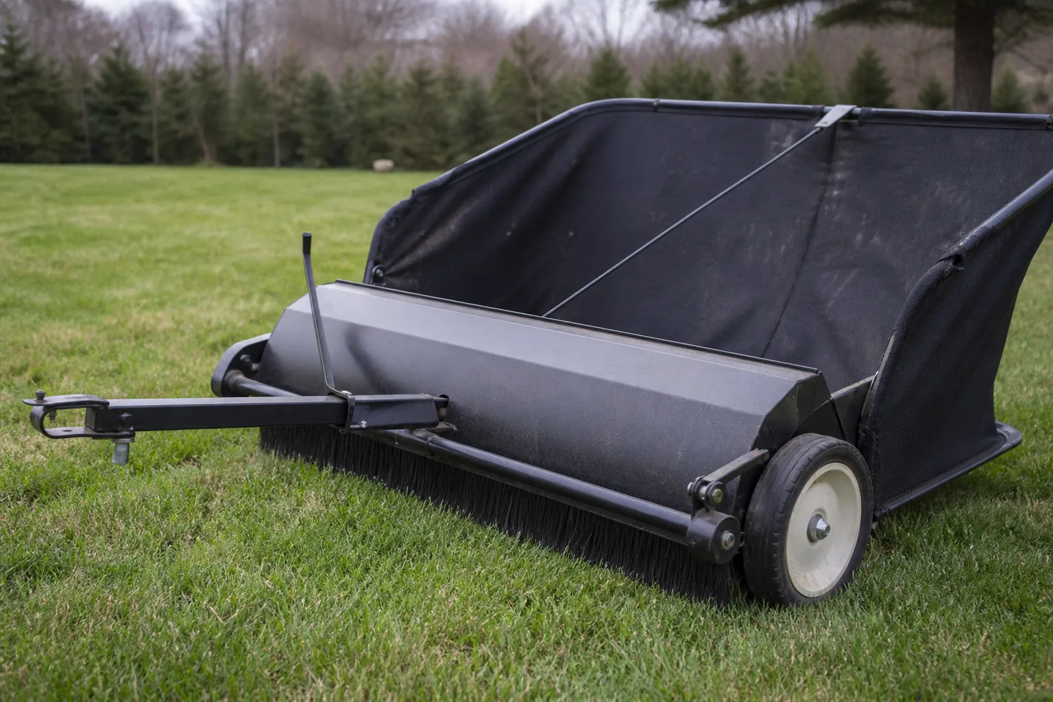 Tow-behind lawn sweeper showing the drawbar hitch, wheel, housing, and brushes touching short grass